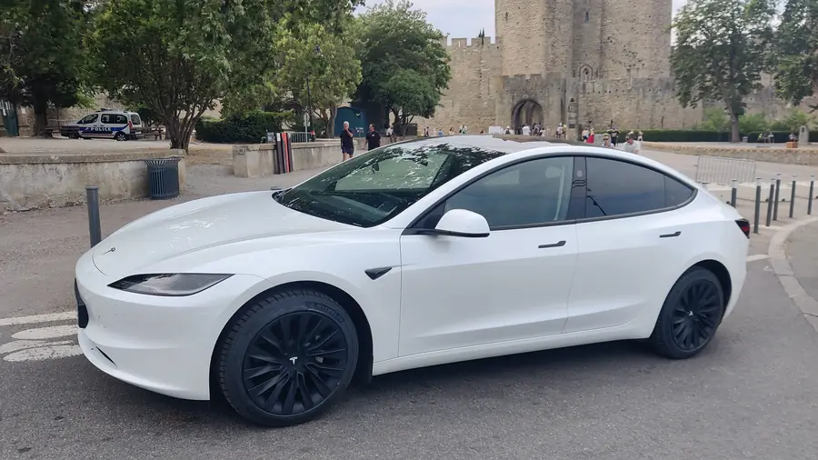 La berline Tesla blanche d’Aude VTC stationnée devant les remparts et les tours d’entrée de la Cité de Carcassonne