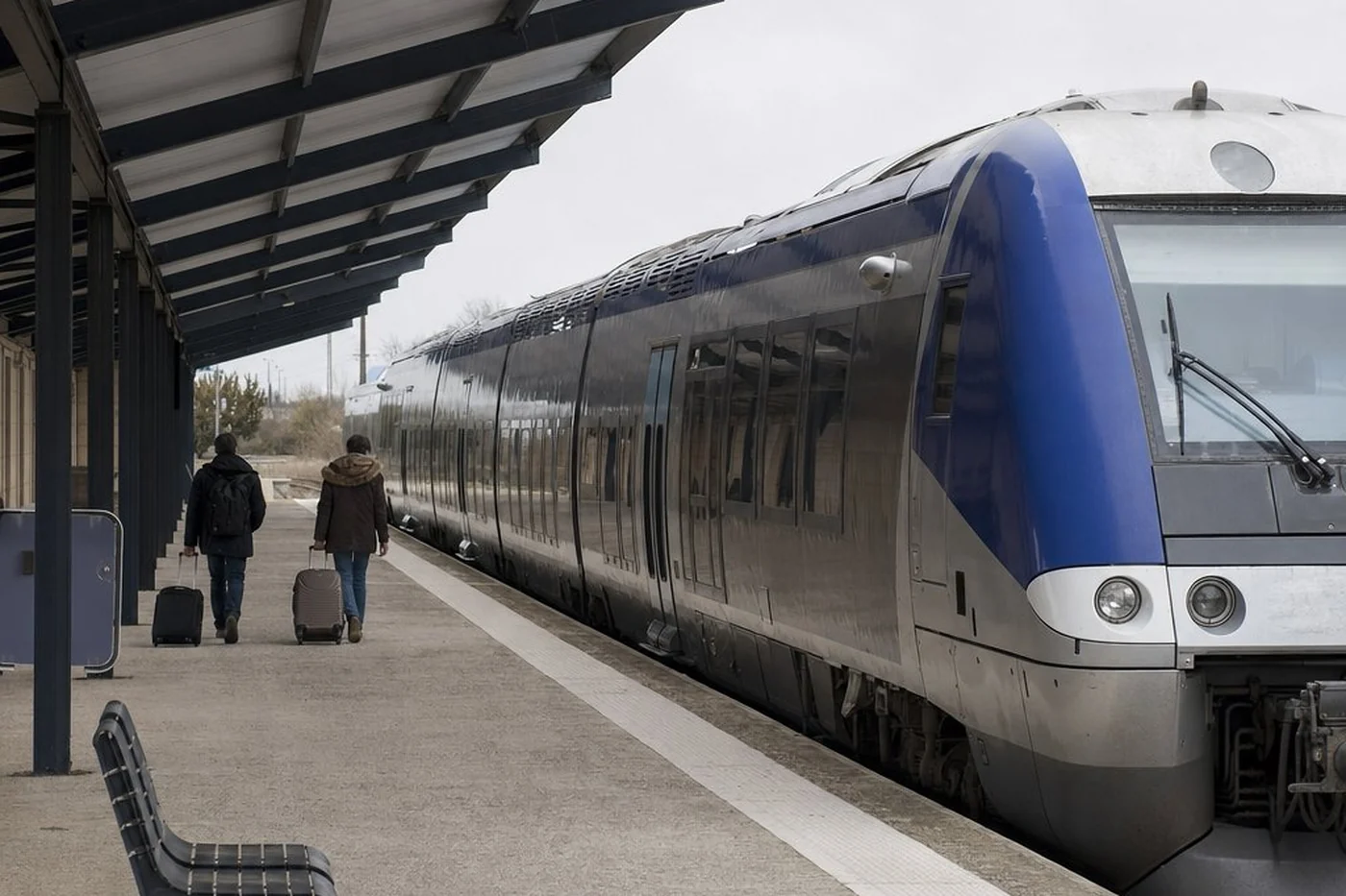 Un train régional à quai dans une petite gare française, deux voyageurs avec des valises à roulettes s’éloignant sur le quai