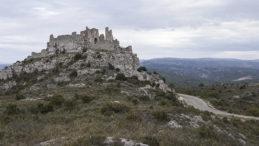 Ruines d’une forteresse médiévale posée sur une crête calcaire couverte de garrigue, une petite route départementale serpentant en contrebas, sous un ciel couvert