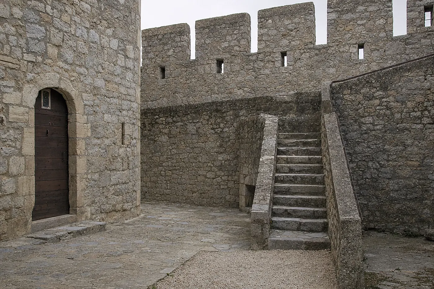 Cour intérieure d’un château médiéval en pierre restauré, escalier de pierre montant vers le chemin de ronde et porte de bois cloutée, sous un ciel couvert